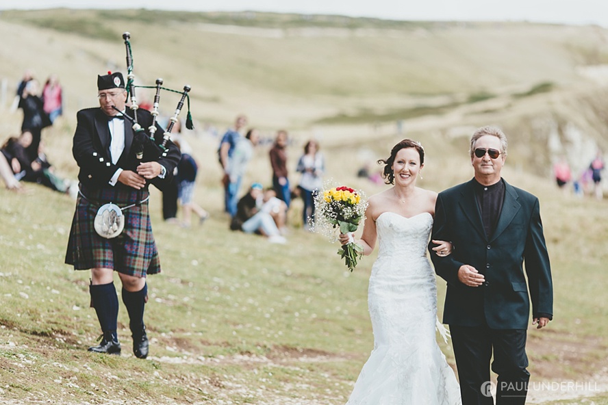 Bride arrives with bagpiper at Durdle Door wedding